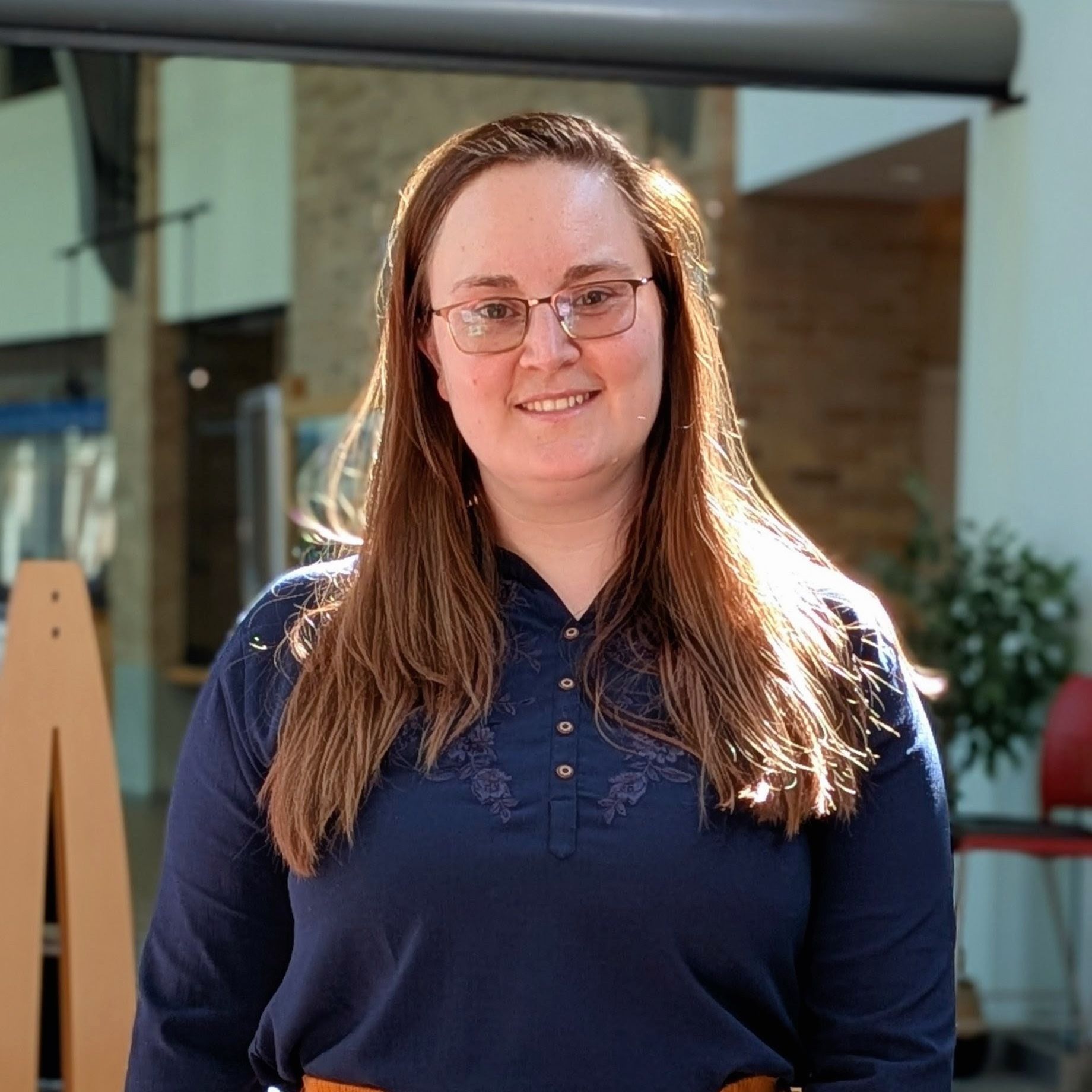 Jeanine Longley at the Port Moody library, wearing a blue shirt and glasses, with her hair down and the sunshine streaming down.